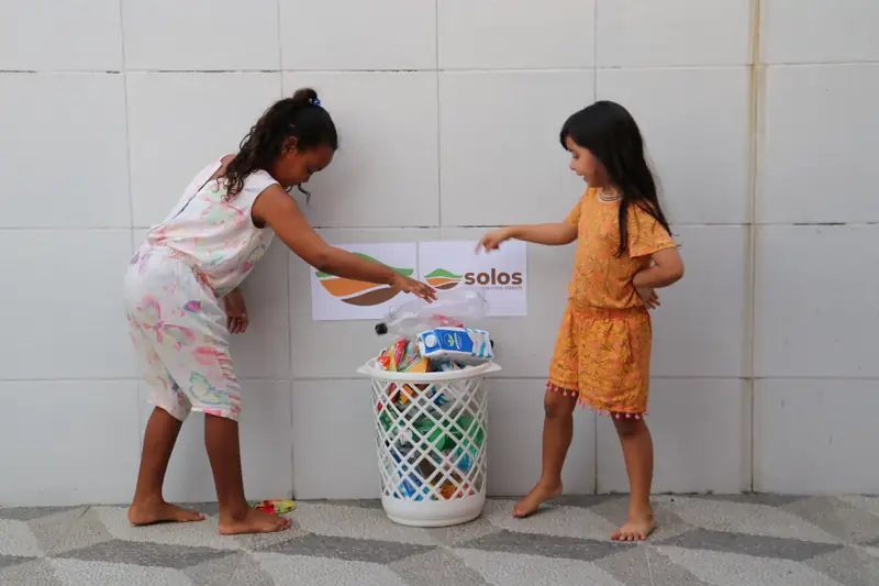 Duas crianças depositando resíduos em uma lixeira com cartaz da Solos