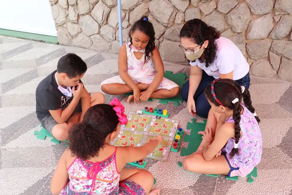 Crianças e educadoras em atividade lúdica com jogo educativo no chão